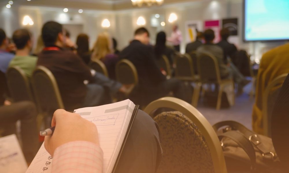 A person is taking notes in a notebook during a conference or seminar, with an audience seated and listening to a speaker at the front of the room, which is illuminated with warm lighting. Lubrication Program