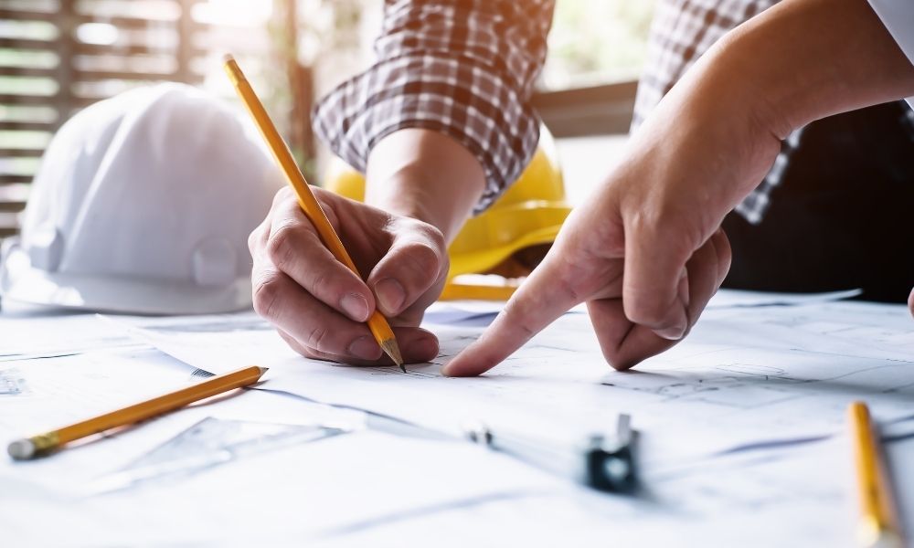 A person in a checkered shirt is pointing at a blueprint or technical drawing on a table, while another person is holding a yellow pencil and making notes or markings. A white safety helmet and a yellow hard hat are on the table, indicating an engineering or construction setting. The scene is well-lit with natural light coming through a window.