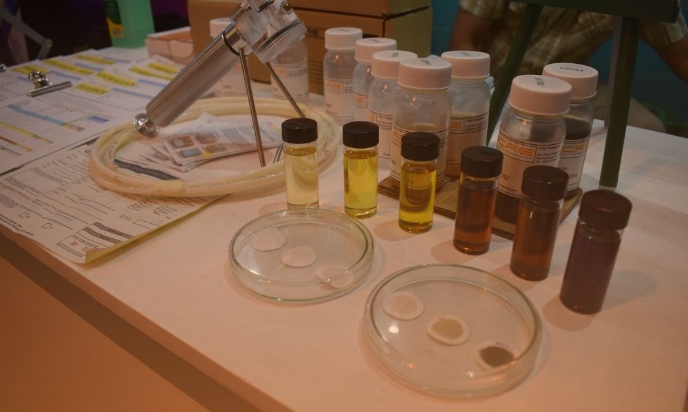 A laboratory workspace featuring several small bottles filled with various colored liquids, some with black caps, placed on a table. There are petri dishes with circular samples, a microscope, and various papers and documents, indicating a scientific or medical testing environment.