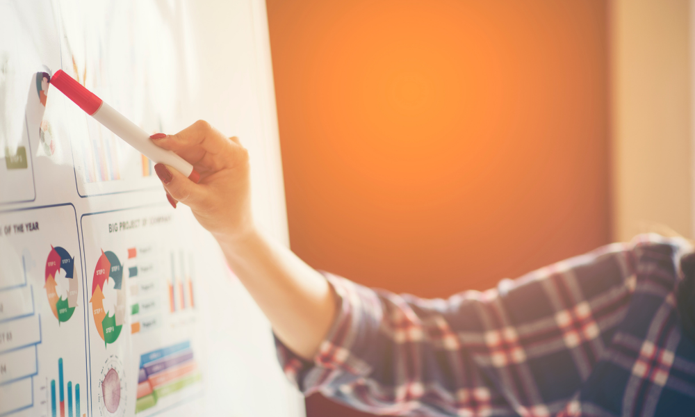A person wearing a plaid shirt is pointing a red and white marker at a whiteboard filled with colorful charts and graphs. The background has warm orange tones, and the focus is on the person's hand and the whiteboard.