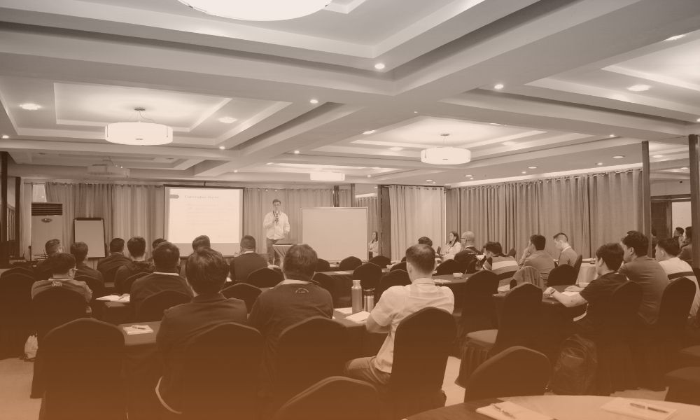 A sepia-toned image of a conference or seminar room filled with attendees seated at tables, facing a presenter standing near a projection screen and whiteboard at the front. The room has a modern ceiling design with recessed lighting and curtains along the walls.