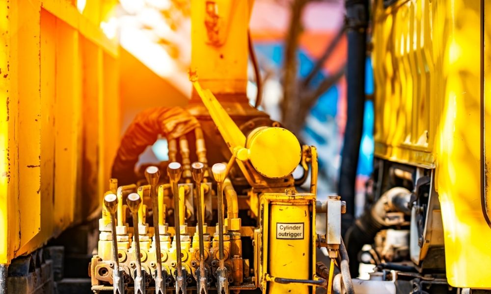 A close-up view of a yellow industrial machine with multiple levers and a labeled cylinder that reads "Cylinder outrigger." The machine appears to be part of heavy construction or manufacturing equipment, with various pipes, hoses, and mechanical components visible. The background includes a mix of blue and orange hues, suggesting an outdoor setting with sunlight.
