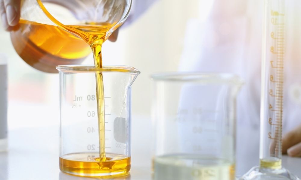 A person pouring a yellow-orange liquid from a beaker into a graduated measuring cylinder in a laboratory setting, with other glassware and scientific equipment visible in the background.