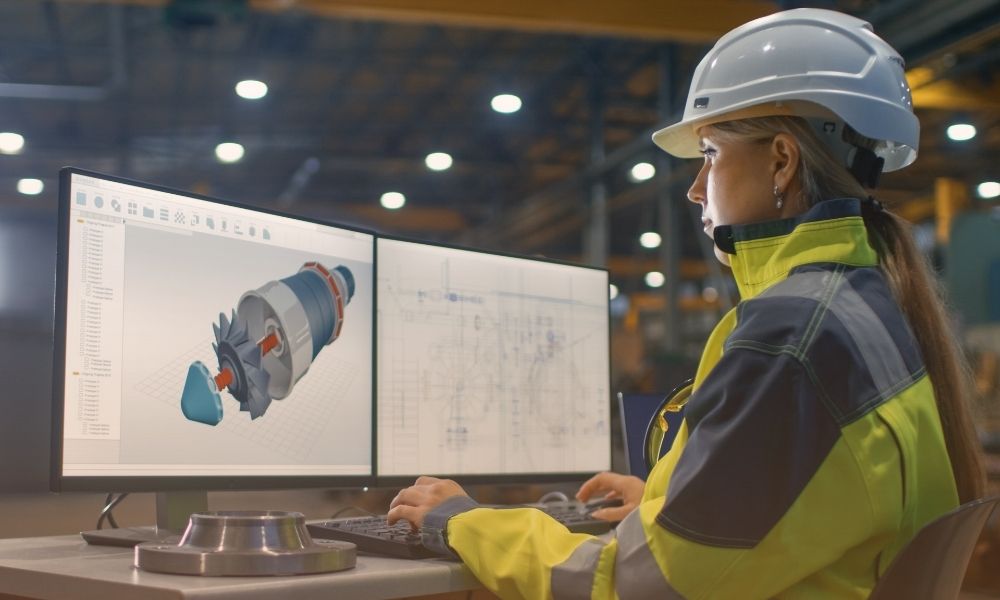 A woman wearing a safety helmet and high-visibility jacket is working at a desk in an industrial setting. She is looking at two large computer monitors displaying technical 3D models and engineering drawings of a mechanical component. The background shows a factory or manufacturing environment with bright overhead lighting.