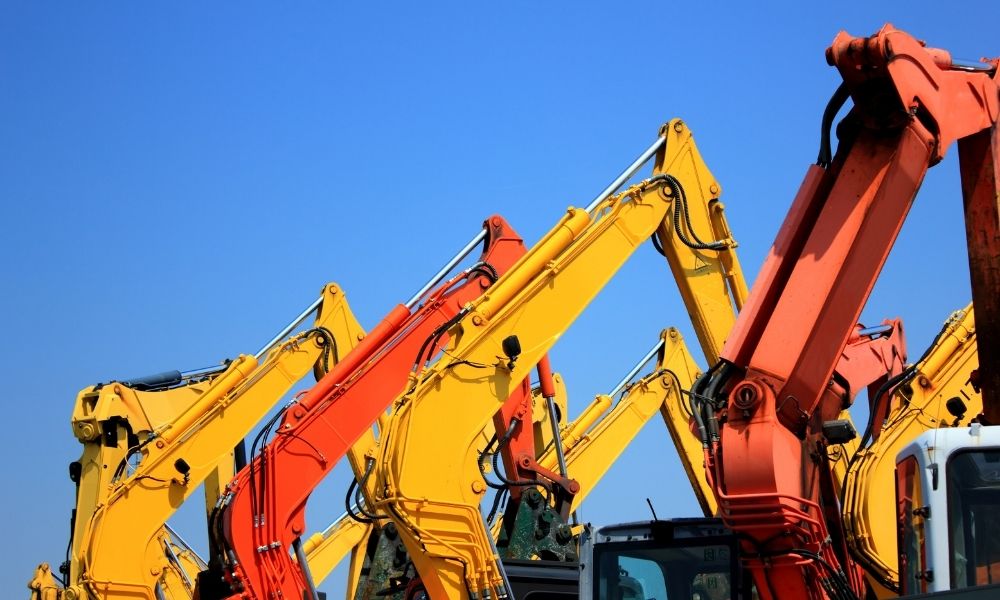 A row of construction excavators with yellow and red arms is set against a clear blue sky, showcasing the machinery lined up closely together.