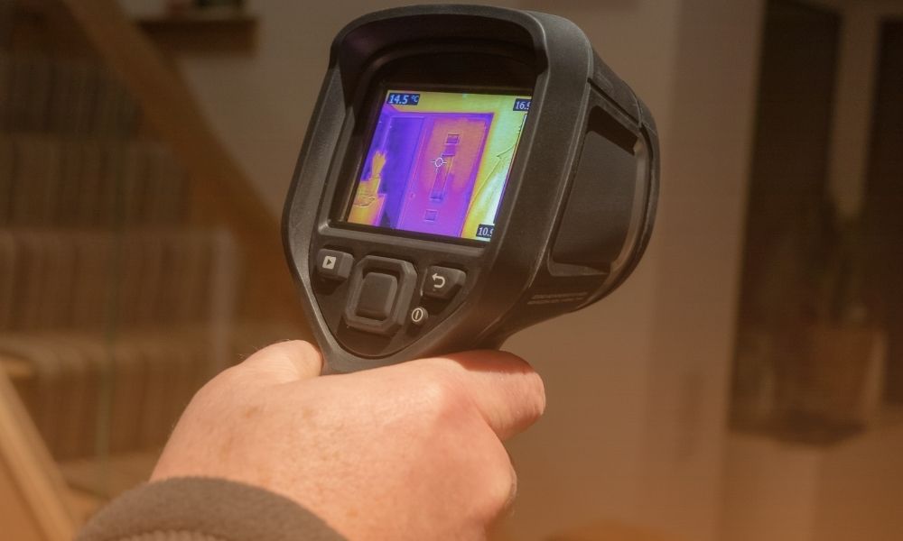 A person holding a thermal imaging camera, which is displaying a thermal image of a room with various objects, including a door and a wall socket. The camera's screen shows different temperature readings in color.