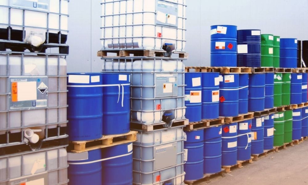 A storage area with numerous large industrial containers and barrels, mostly blue, green, and gray, stacked on wooden pallets against a plain wall.
