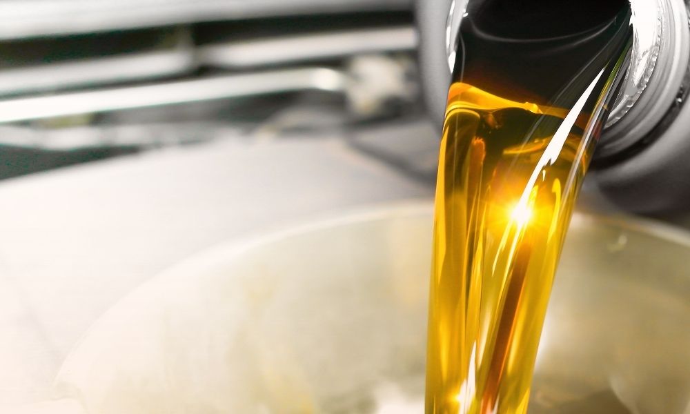 Golden motor oil is being poured into a container, with the oil flowing smoothly and reflecting light. The background is blurred, focusing attention on the rich, amber-colored oil.