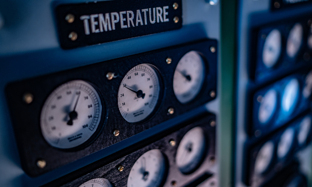 A close-up of an industrial control panel with multiple gauges and meters, including a prominent temperature gauge at the top left. The panel has a label that reads "TEMPERATURE" above the gauges, and the gauges display various readings with needle indicators. The overall setting appears to be in a control room or machinery environment.