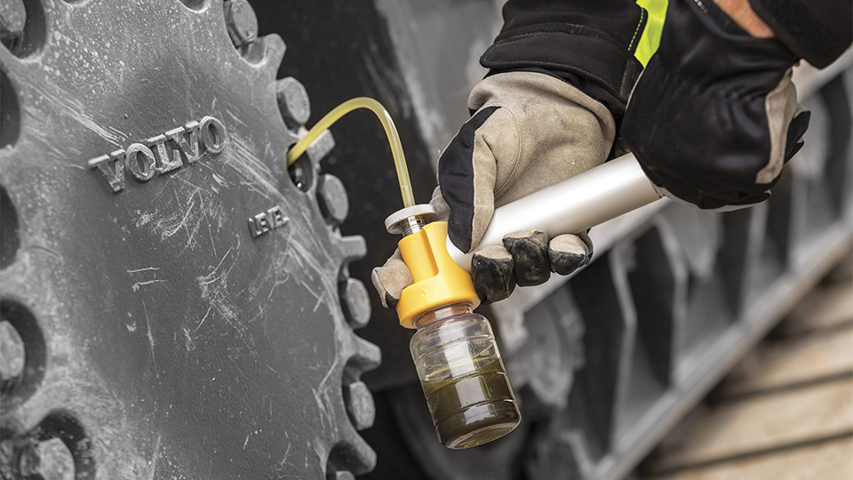 A person wearing gloves is holding a small container connected to a syringe, extracting Oil Sampling or injecting dark liquid near the large gear of a Volvo machine, indicating maintenance or fluid testing. (Oil Sampling Hardware Guide )