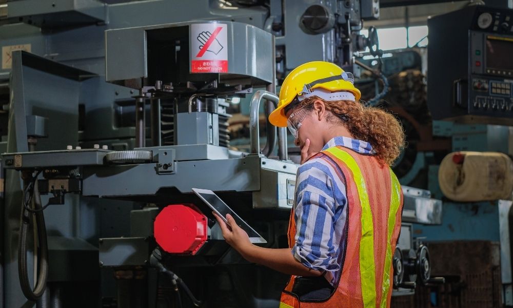 A female industrial worker wears a yellow safety helmet, safety glasses, and a reflective orange vest as she inspects or operates a large piece of machinery in an industrial setting. She holds a tablet device, her eyes fixed on the screen as she scrolls through images of wear debris on the equipment's mechanical components. The background is a maze of control panels and machinery parts, with a few loose screws and scraps of metal scattered about.