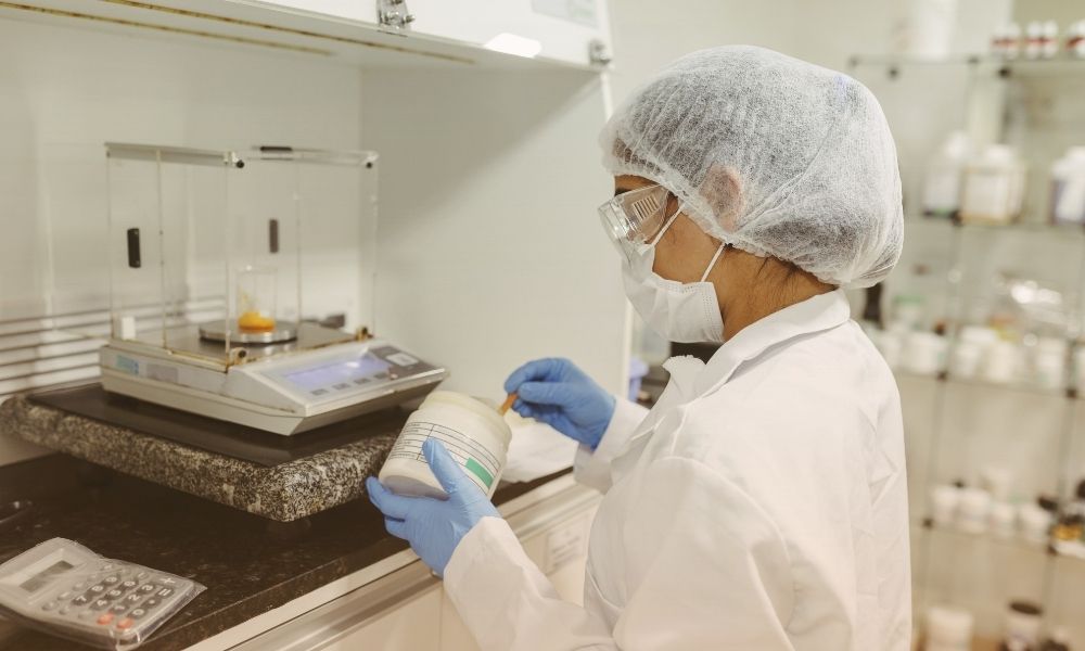 A scientist or laboratory technician wearing a hairnet, face mask, safety glasses, and gloves is working in a laboratory. They are holding a container and appear to be taking notes or recording information, with a balance scale and other lab equipment in the background.