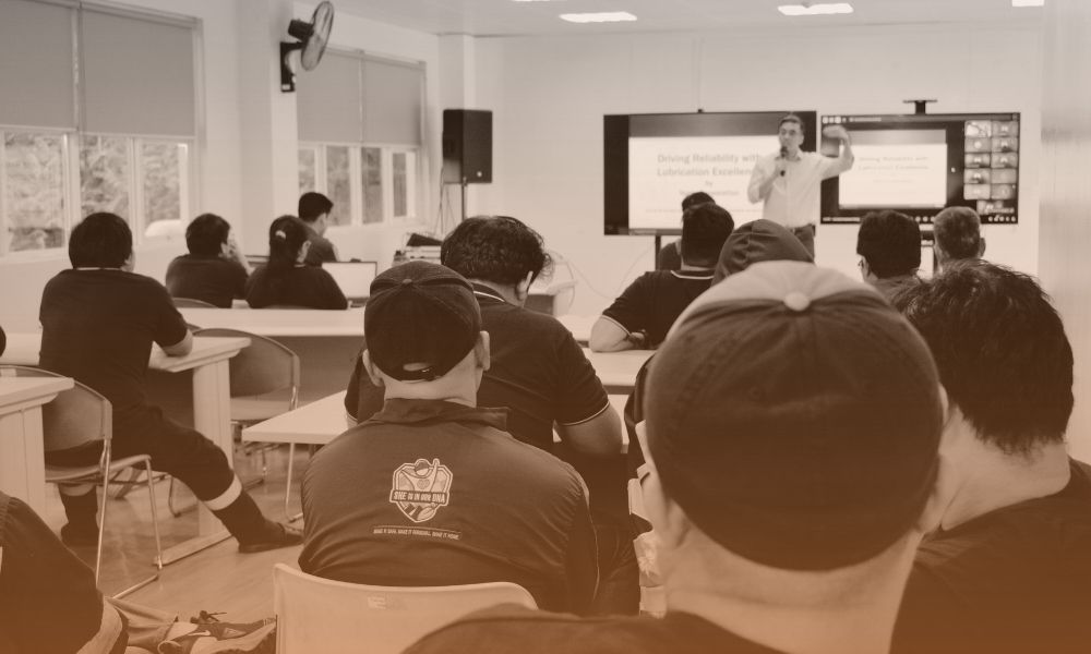 A classroom or training room with students seated at desks, attentively listening to a presenter at the front. The presenter is standing near a large screen displaying a presentation slide, and appears to be explaining something, gesturing with his hand. The room has multiple windows, and the overall tone of the image is sepia.