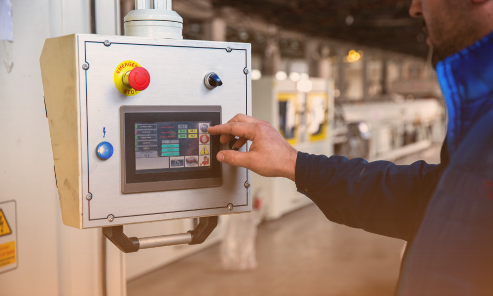A person operating an industrial control panel with a touchscreen interface in a factory or manufacturing environment. The panel has emergency stop and other control buttons, and the person is interacting with the touchscreen to monitor or adjust machinery.
