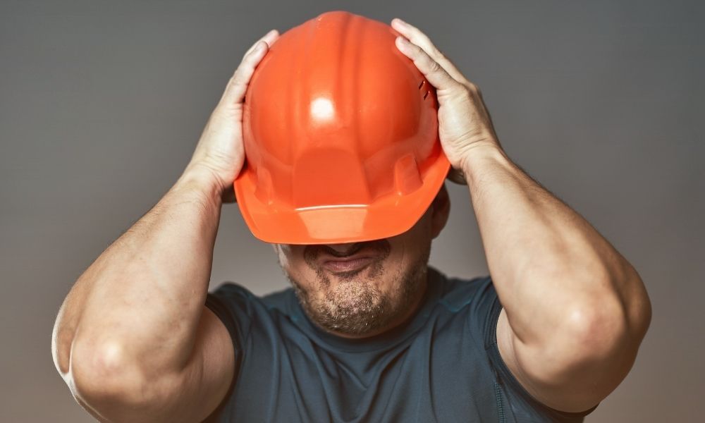 A man wearing a blue shirt is holding an orange safety helmet over his head with both hands, partially covering his face. The background is plain and gray.