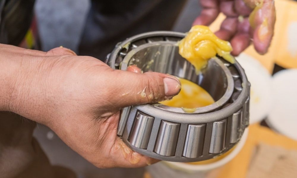 An individual is holding a large ball bearing with their left hand, while their right hand is placing or removing a yellowish substance, possibly grease, onto or from the bearing. The background includes blurred objects, suggesting a workshop or mechanical setting.