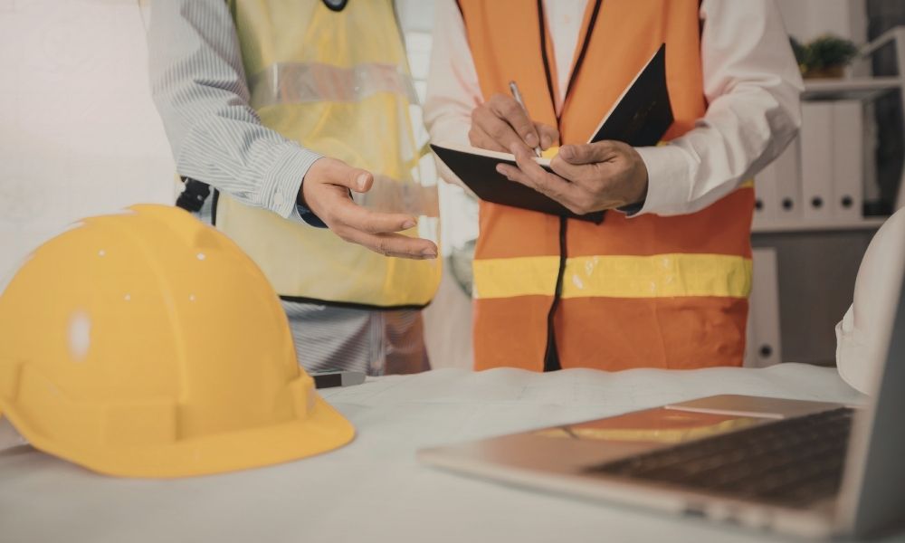 A construction supervisor and a worker wearing safety vests are having a discussion in an office or planning area. The supervisor is holding a notebook and pen, taking notes, while the worker gestures with their hand. A yellow safety helmet is placed on the table in front of them. In the background, there are shelves with binders and office supplies.