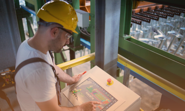 A worker wearing a yellow safety helmet and safety glasses operating a control panel with a touchscreen interface, in an industrial setting with machinery and green structural components around him.