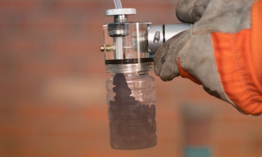 A person wearing gloves is using a tool to extract or insert a plastic bottle filled with black charcoal or a similar material, with a brick wall in the background.