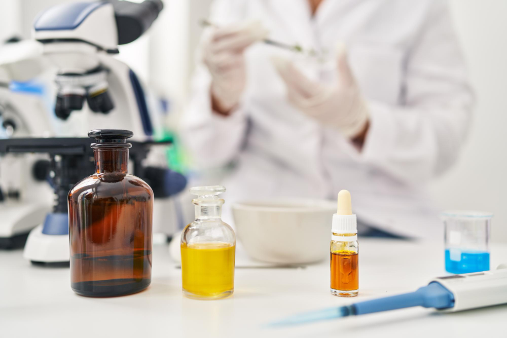 An out-of-focus scientist in a white lab coat and gloves works in the background of a bright laboratory. In the sharp foreground, three glass bottles containing different oils or liquids sit on a white table: a large amber apothecary bottle, a small clear bottle with a bright yellow liquid, and a tiny dropper bottle with dark orange oil. A microscope, a mortar and pestle, a small beaker with blue liquid, and a pipette are also visible on the workspace.