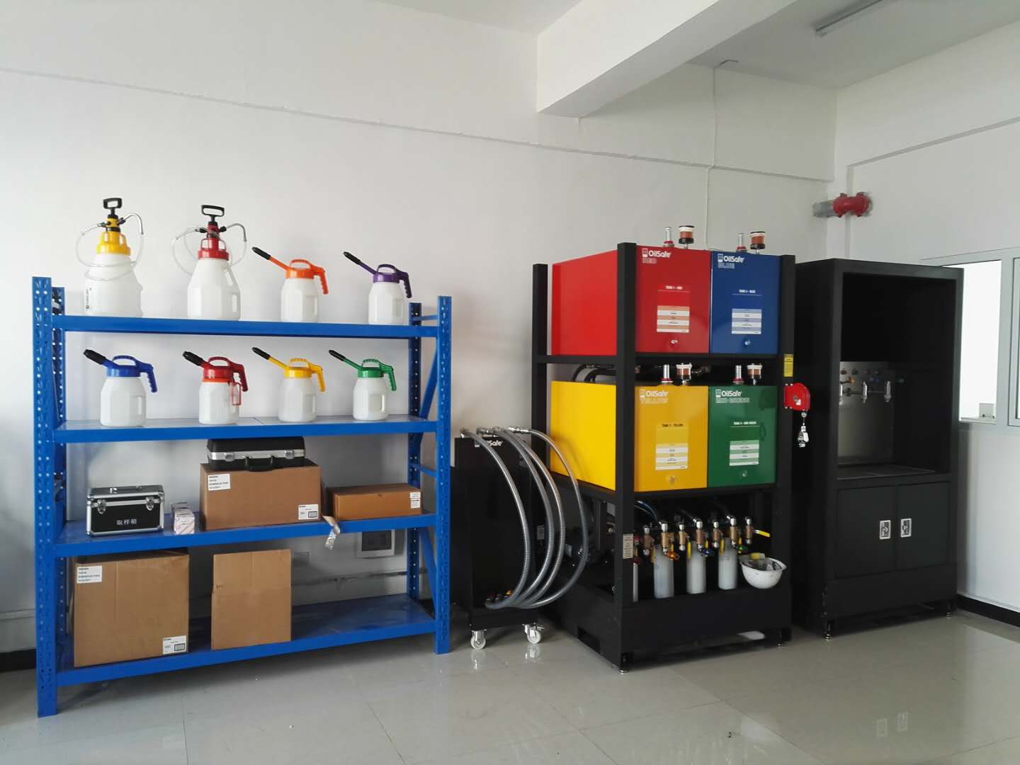 A laboratory corner with a blue metal shelf holding several spray bottles with different colored nozzles, and a black and brown box on the bottom shelf. Next to the shelf is a black equipment rack with colorful containers and hoses connected to various devices, including a water or chemical dispensing system. The background features a plain white wall, a window, and a ceiling with fluorescent lighting.