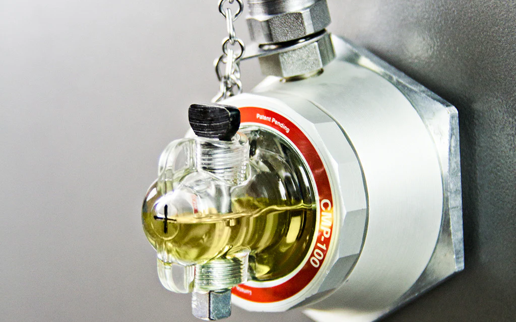 An industrial device with a transparent glass component filled with a yellowish liquid, attached to a metallic fixture. The device features a red and white circular label reading "CAMP-100" and "Patent Pending." The fixture is mounted on a gray wall, with a chain and a screw mechanism visible at the top.