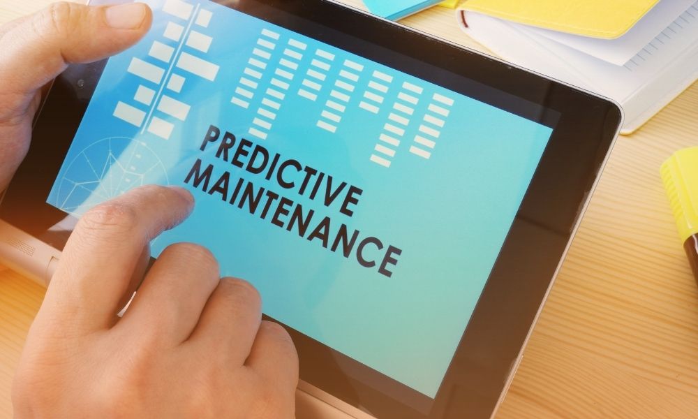 A person is touching a tablet screen displaying the words "PREDICTIVE MAINTENANCE" along with various graphical elements, on a desk with yellow highlighters, notebooks, and papers around it.