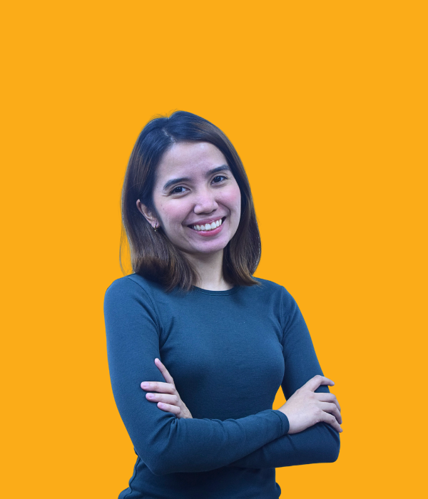 A young woman with shoulder-length brown hair, smiling confidently with her arms crossed, wearing a long-sleeved dark blue top against an orange background.