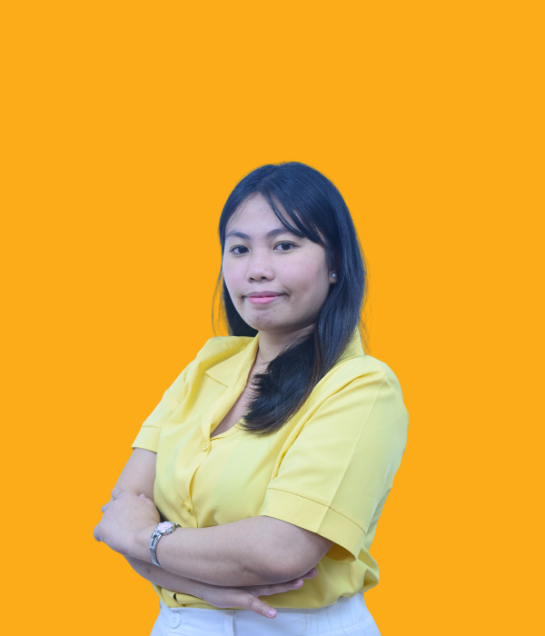 A young woman with shoulder-length dark hair, wearing a yellow blouse and white pants, standing with her arms crossed against an orange background. She has a confident expression on her face.