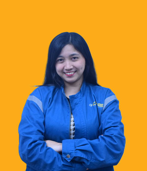 A young woman with shoulder-length black hair, smiling and standing with her arms crossed, wearing a blue jacket with a logo on the chest, against a solid orange background.