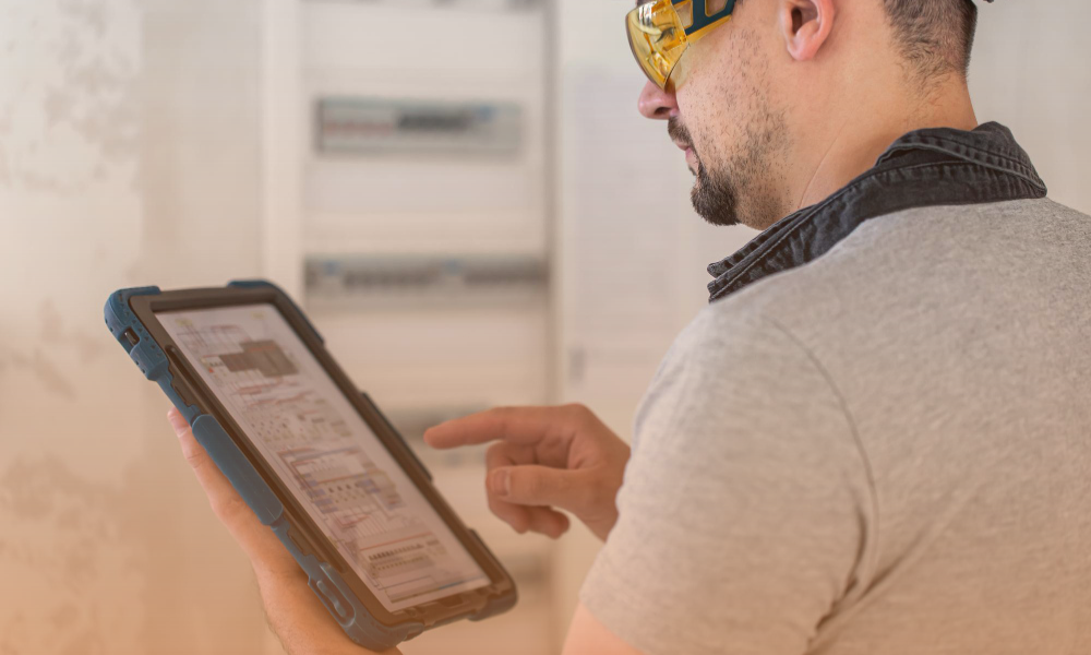 A man wearing safety glasses and a gray shirt is holding and interacting with a tablet device, likely inspecting or analyzing data in an industrial or technical environment. The background shows electrical panels or equipment.