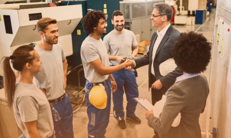 A group of five workers, including women and men, are in an industrial or manufacturing plant. They are smiling and engaging in conversation or a handshake with a suited man, possibly a manager or visitor. One worker is holding a yellow safety helmet, and another person is holding a clipboard. The background features machinery and industrial equipment.