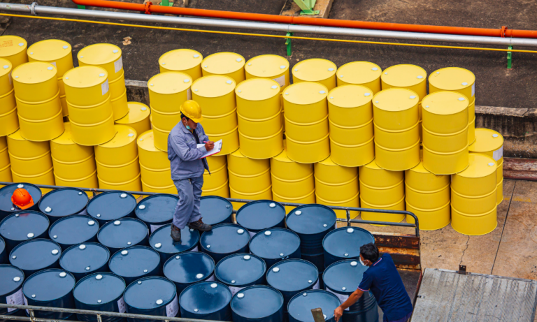 A worker wearing a yellow safety helmet and blue uniform standing among yellow and black oil drums, taking notes on a clipboard.